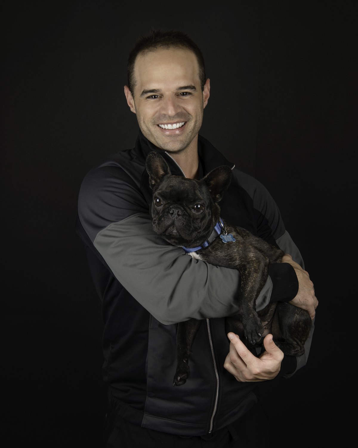 Dr. Jeff Trembley smiling and holding Peewee, the studio's French Bulldog, against a black backdrop